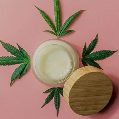 An Open Jar Of Medical Cannabis Topical Cream On Table With Bamboo Lid Encircled By Four Cannabis Leaves - Topicals Category Background Cover Image